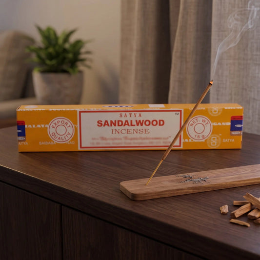 Satya Sandalwood Räucherstäbchen: glimmendes Räucherstäbchen mit feinem Rauch im Holzhalter, dahinter die Packung, auf einem Sideboard am Abend