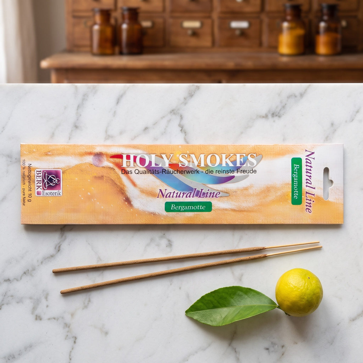 Bergamotte Räucherstäbchen der Holy Smokes Natural Line von Berk: flache Packung auf einer Marmortheke vor einem alten Holzschrank mit Schubladen und Bernsteinglasflaschen, davor zwei lose Stäbchen und eine Bergamotte mit Blatt