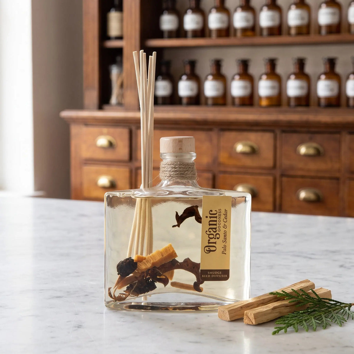 Reed Diffuser Palo Santo und Zeder von Organic Goodness: eckige Glasflasche mit Holzstopfen, Jutekordel und Schilfrohrstäbchen auf einer Marmortheke vor einem Apothekerschrank