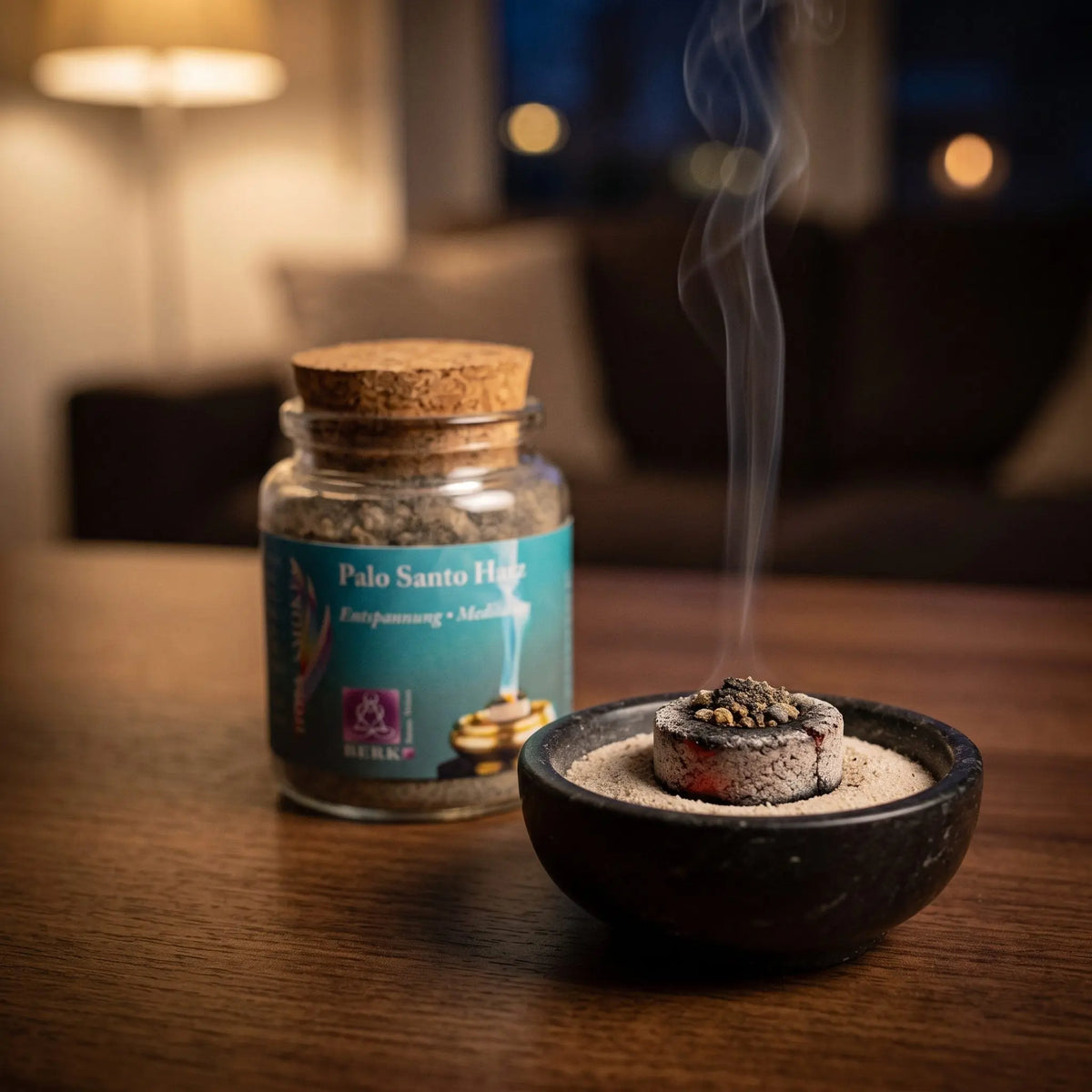 Palo Santo Harz von Berk, 60 ml im Korkglas: glimmende Räucherkohle mit einer Prise Palo-Santo-Harz in einer Specksteinschale, dahinter das Korkglas, feiner Rauch steigt auf einer dunklen Holzplatte am Abend auf – Spirit of Gaia