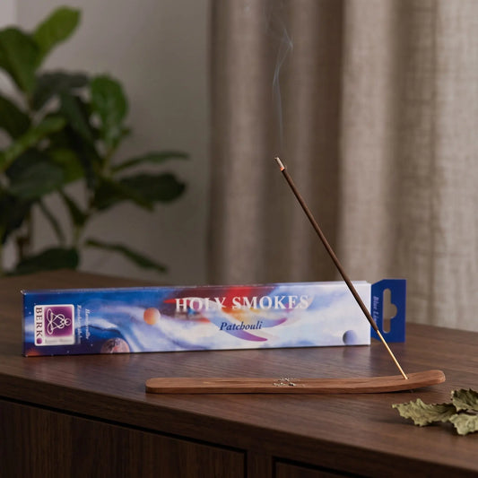 Patchouli Räucherstäbchen Holy Smokes Blue Line: glimmendes Räucherstäbchen mit feinem Rauch im Holzhalter, dahinter die Packung, auf einem Sideboard am Abend