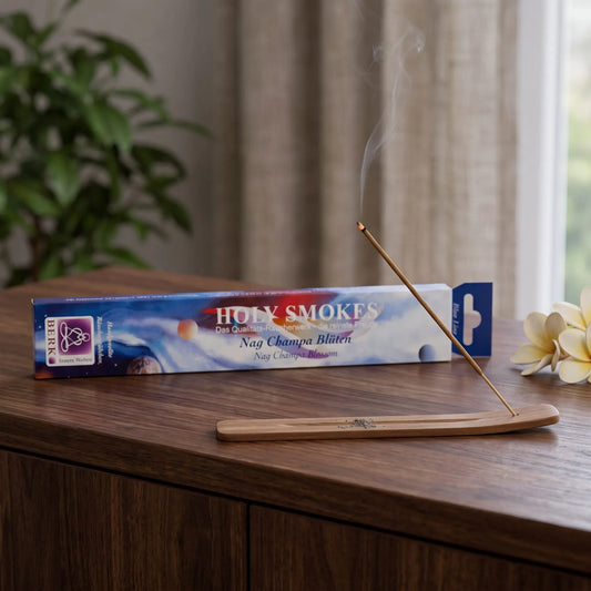 Nag Champa Blüten Räucherstäbchen Blue Line: glimmendes Räucherstäbchen mit feinem Rauch im Holzhalter, dahinter die Packung, auf einem Sideboard am Abend
