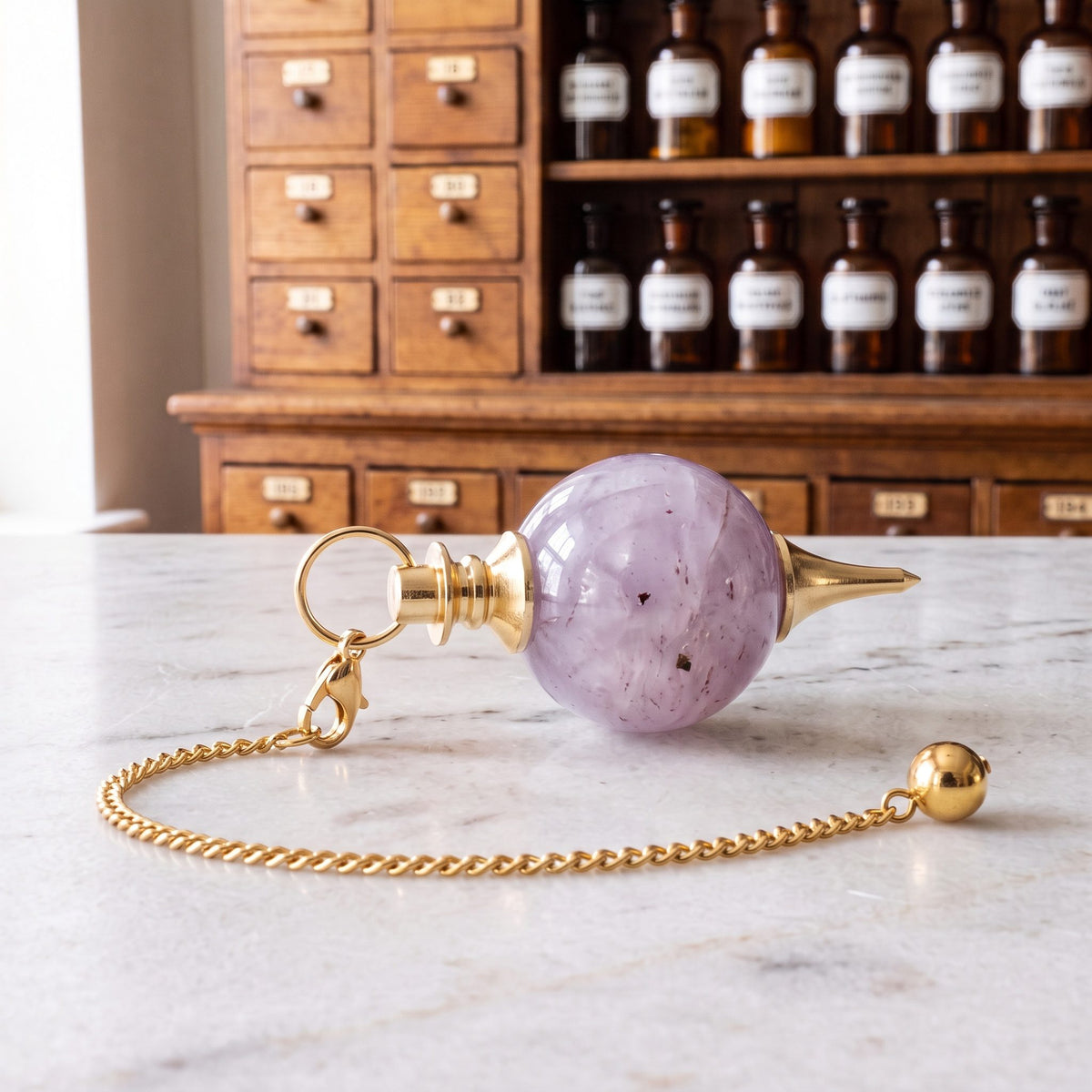 Amethyst-Kugelpendel von Berk liegt auf der Marmortheke einer hellen Apotheke, die goldfarbene Kette mit Karabiner und Endkugel locker daneben