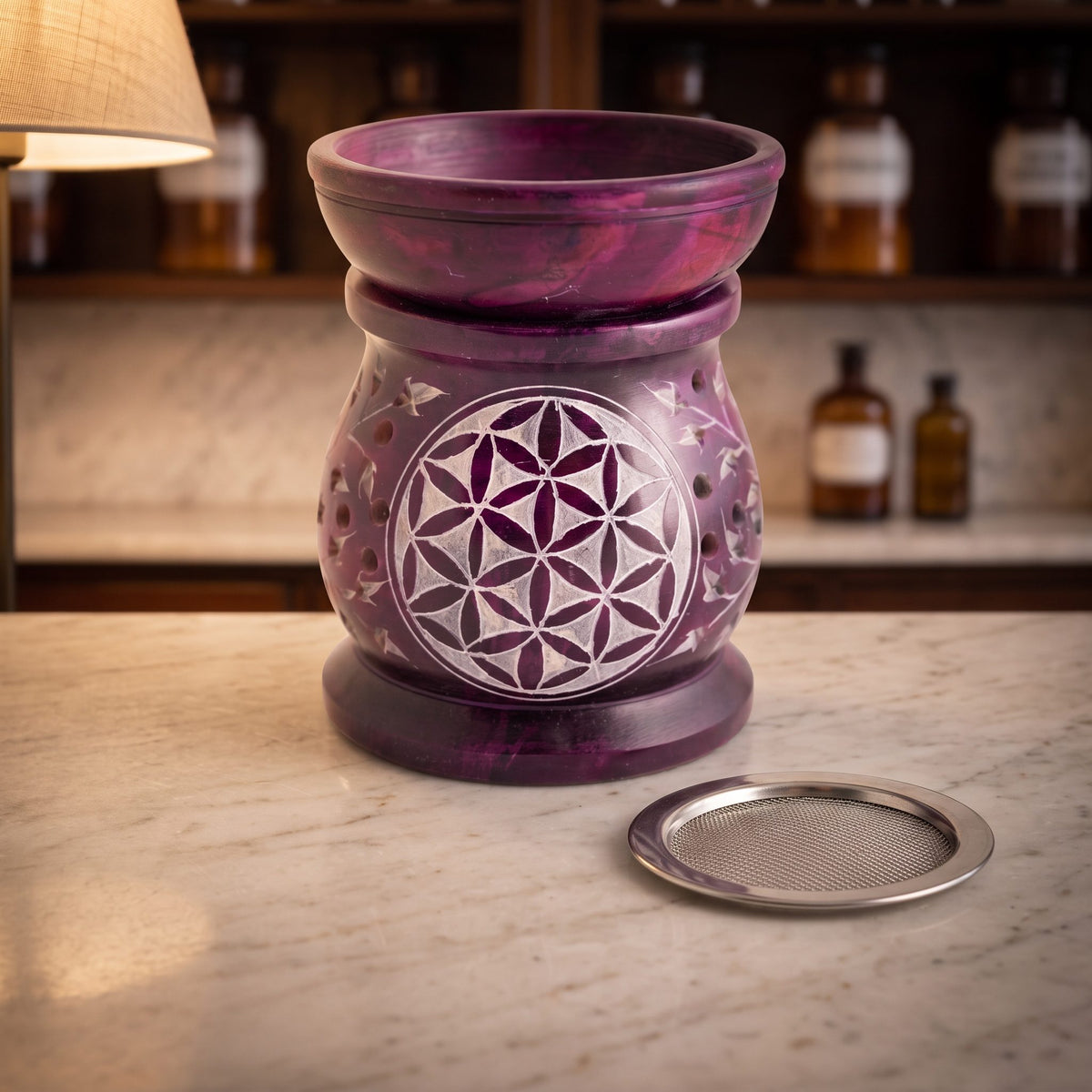 Aromalampe und Siebgefäß Blume des Lebens aus violettem Speckstein mit aufgesetzter Schale, davor das Edelstahlsieb, auf der Marmortheke einer Apotheke am Abend