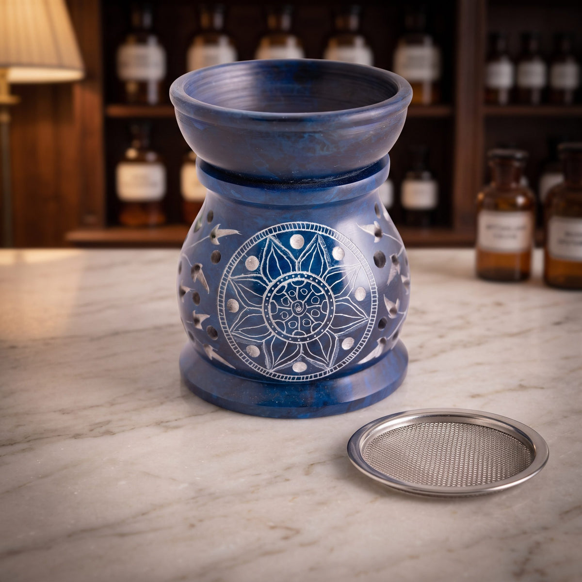Aromalampe und Siebgefäß Mandala aus blauem Speckstein mit aufgesetzter Schale, davor das Edelstahlsieb, auf der Marmortheke einer Apotheke am Abend
