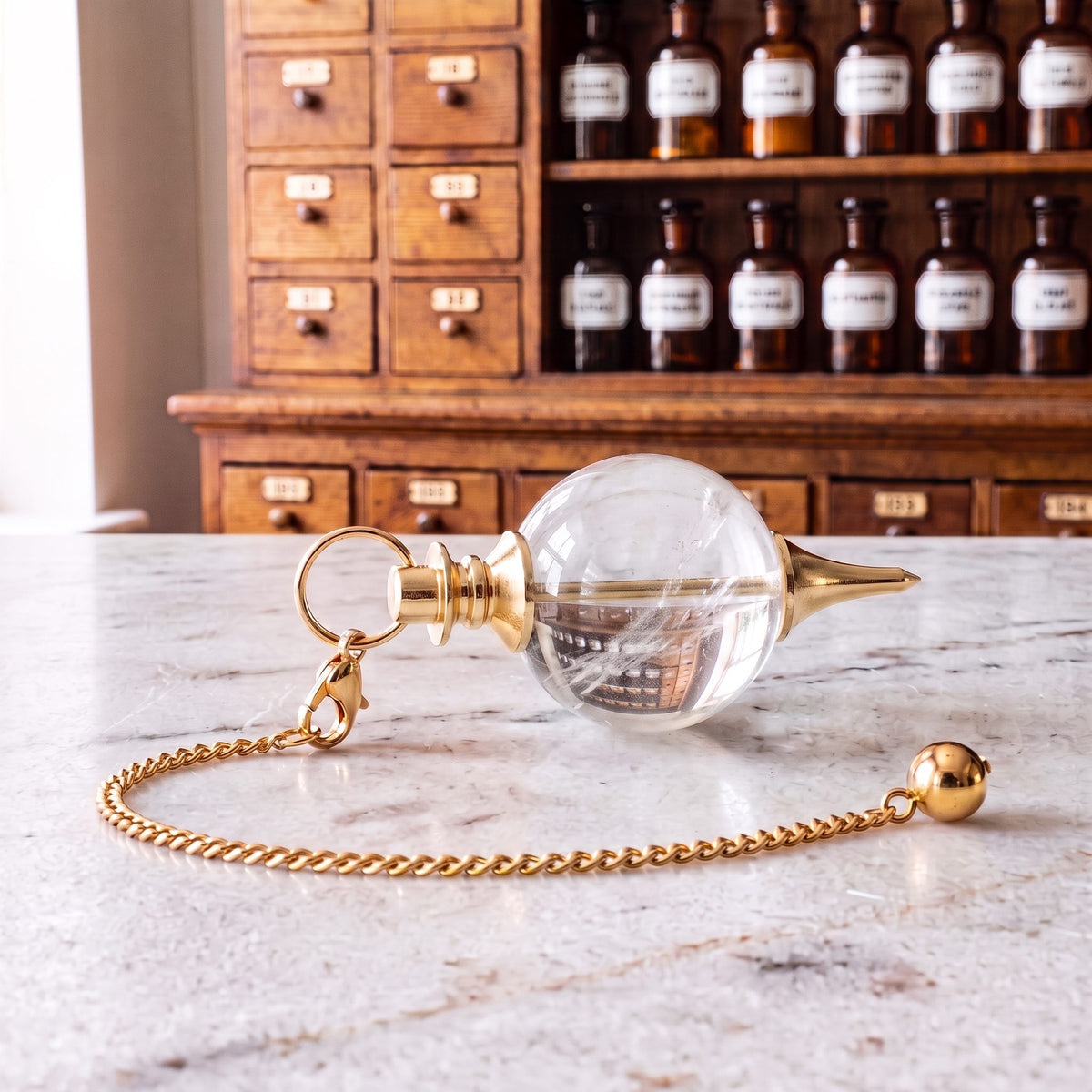 Bergkristall-Kugelpendel von Berk liegt auf der Marmortheke einer hellen Apotheke, die goldfarbene Kette mit Karabiner und Endkugel locker daneben
