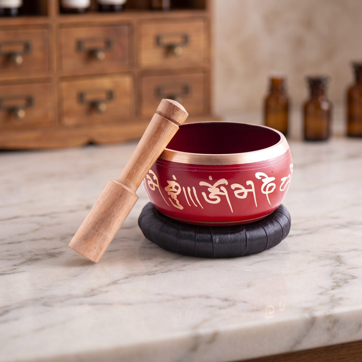 Rote Klangschale mit goldenem Rand und goldenem Mantra-Schriftband auf schwarzer Unterlage mit Klangstab auf einer Marmortheke