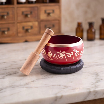 Rote Klangschale mit goldenem Rand und goldenem Mantra-Schriftband auf schwarzer Unterlage mit Klangstab auf einer Marmortheke