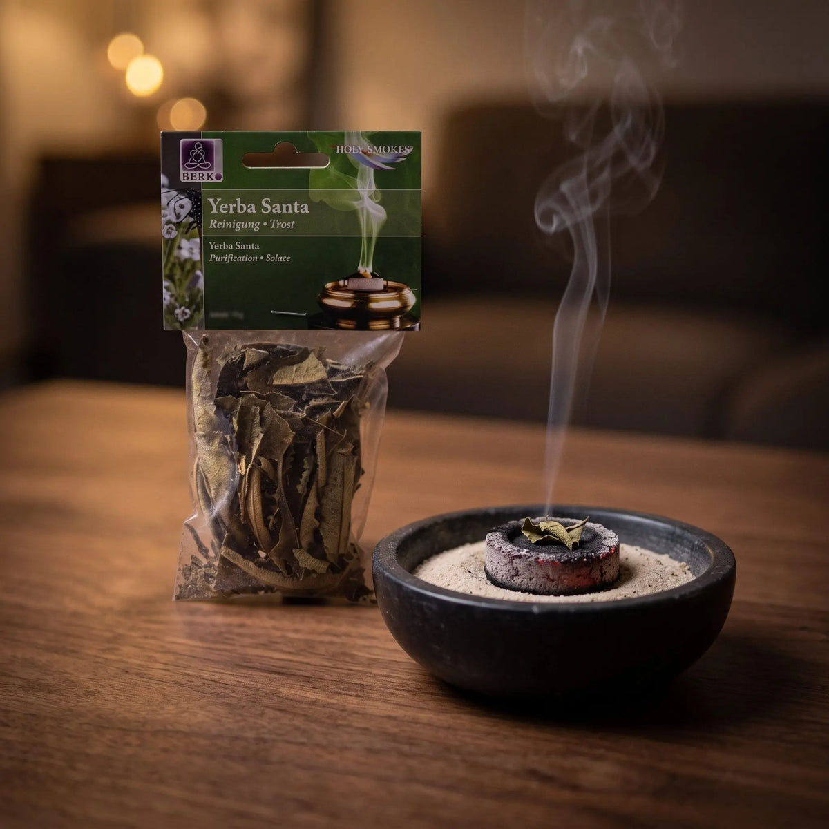 Yerba Santa, sortenreines Räucherwerk von Berk, 10 g: ein Stück Yerba-Santa-Blatt glimmt auf der Räucherkohle, dahinter die Tüte, feiner Rauch steigt auf einer dunklen Holzplatte am Abend auf – Spirit of Gaia