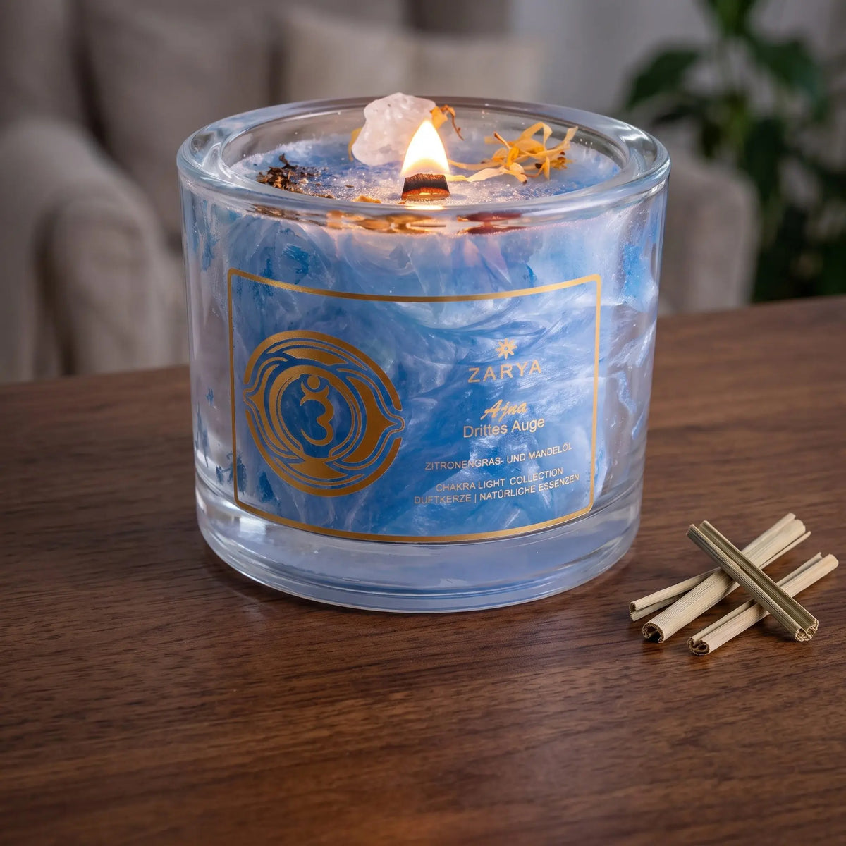 Chakra-Kerze Stirnchakra Ajna mit Bergkristall, Duft Zitronengras und Mandel: brennende Kerze mit kleiner Flamme am Abend auf einer dunklen Holzplatte im Wohnzimmer, daneben getrocknetes Zitronengras – Spirit of Gaia