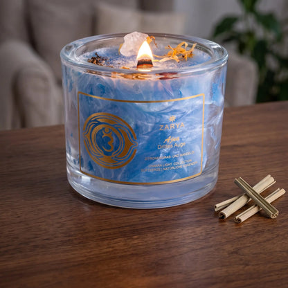 Chakra-Kerze Stirnchakra Ajna mit Bergkristall, Duft Zitronengras und Mandel: brennende Kerze mit kleiner Flamme am Abend auf einer dunklen Holzplatte im Wohnzimmer, daneben getrocknetes Zitronengras – Spirit of Gaia