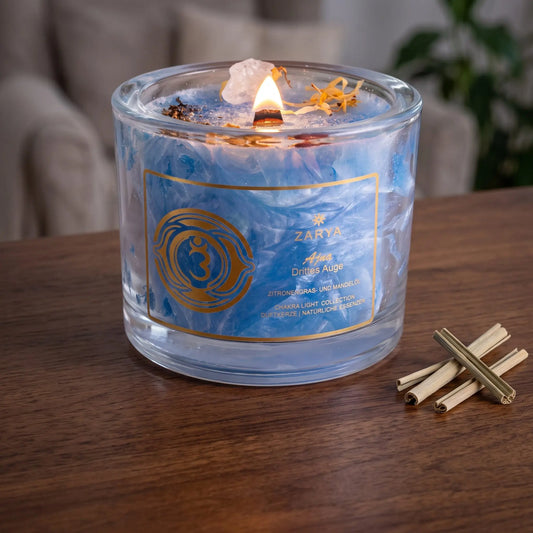 Chakra-Kerze Stirnchakra Ajna mit Bergkristall, Duft Zitronengras und Mandel: brennende Kerze mit kleiner Flamme am Abend auf einer dunklen Holzplatte im Wohnzimmer, daneben getrocknetes Zitronengras – Spirit of Gaia
