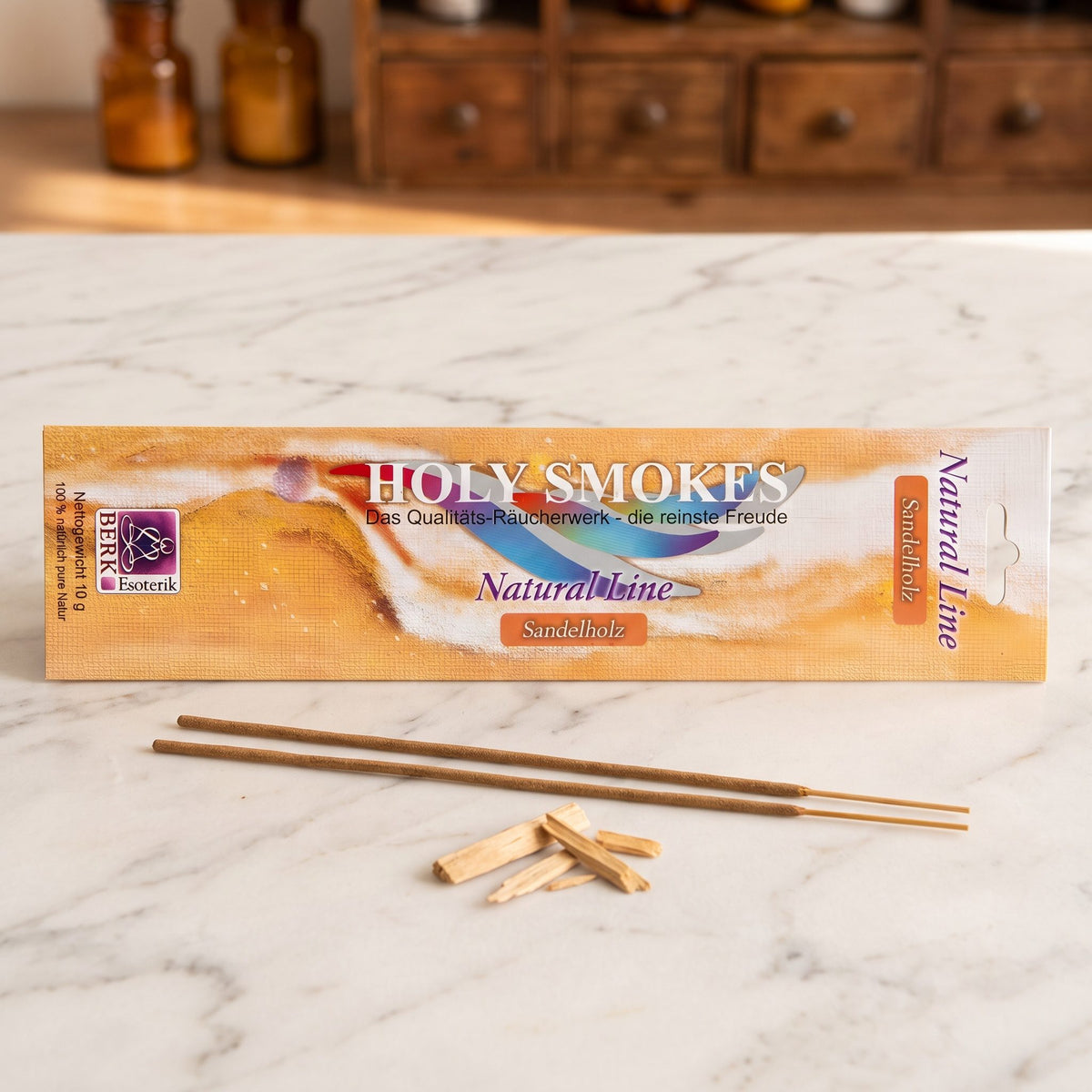 Sandelholz Räucherstäbchen der Holy Smokes Natural Line von Berk: flache Packung auf einer Marmortheke vor einem alten Holzschrank mit Schubladen und Bernsteinglasflaschen, davor zwei lose Stäbchen und Sandelholzspäne