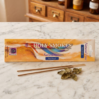 Patchouli Räucherstäbchen der Holy Smokes Natural Line von Berk: flache Packung auf einer Marmortheke vor einem alten Holzschrank mit Schubladen und Bernsteinglasflaschen, davor zwei lose Stäbchen und getrocknete Patchouliblätter