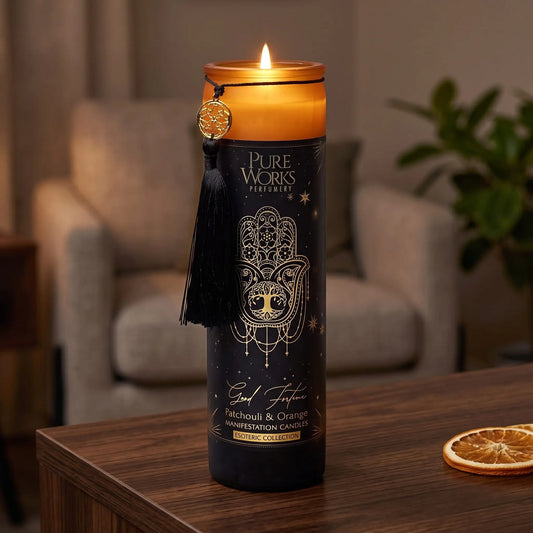 Manifestationskerze Good Fortune (Patchouli & Orange): brennende Glaskerze auf einem dunklen Holztisch im Wohnzimmer am Abend, daneben eine getrocknete Orangenscheibe