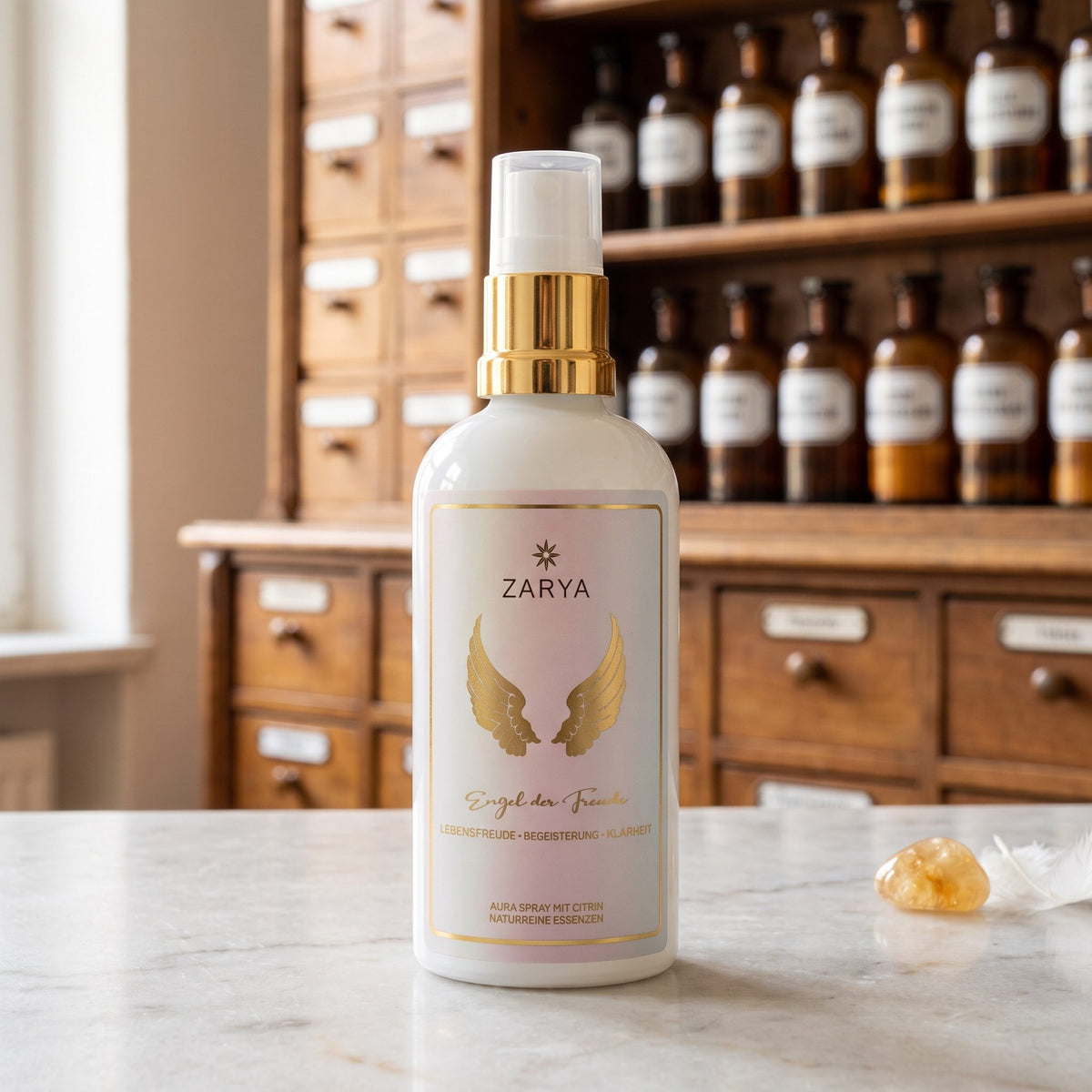Engel der Freude Aura Spray von Zarya: weiße Flasche mit goldenem Pumpring und goldenen Flügeln auf dem Etikett, auf der Marmortheke einer Apotheke, daneben ein gelber Citrin und eine weiße Feder