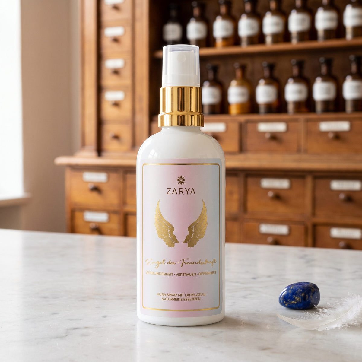 Engel der Freundschaft Aura Spray von Zarya: weiße Flasche mit goldenem Pumpring und goldenen Flügeln auf dem Etikett, auf der Marmortheke einer Apotheke, daneben ein blauer Lapislazuli und eine weiße Feder