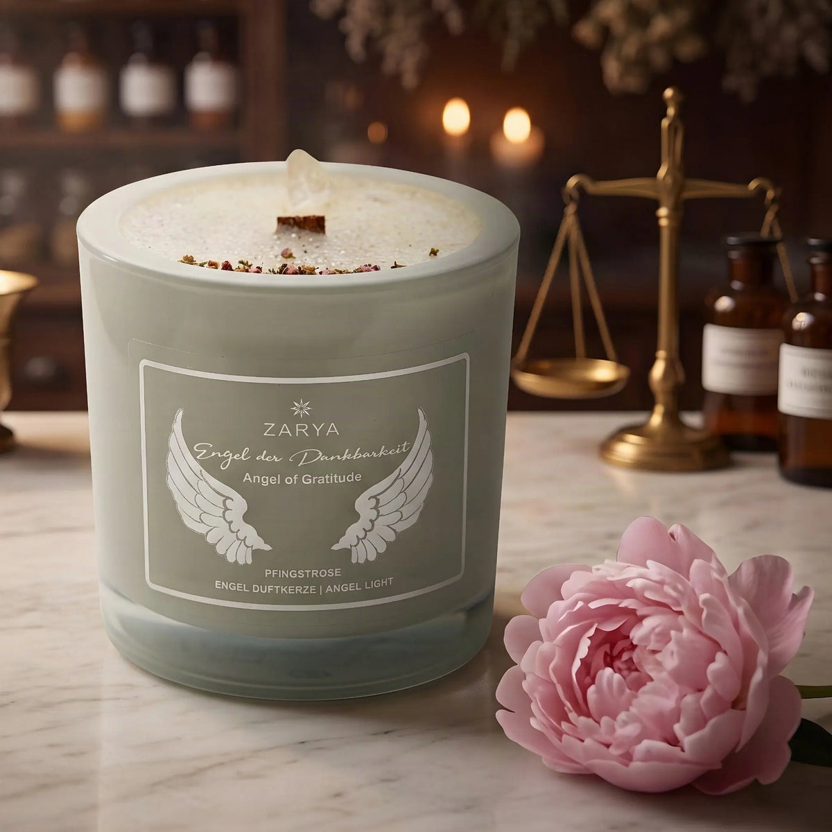 Engelkerze Engel der Dankbarkeit von Zarya: salbeigrünes Glas mit weißem Aufdruck auf der Marmortheke einer alten Apotheke am Abend, daneben eine Pfingstrosenblüte – Spirit of Gaia