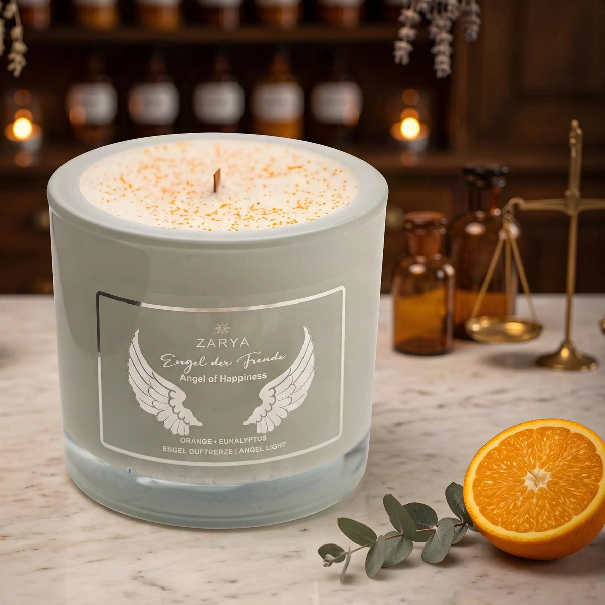 Engelkerze Engel der Freude von Zarya: salbeigrünes Glas mit weißem Aufdruck auf der Marmortheke einer alten Apotheke am Abend, daneben eine halbe Orange und ein Eukalyptuszweig – Spirit of Gaia