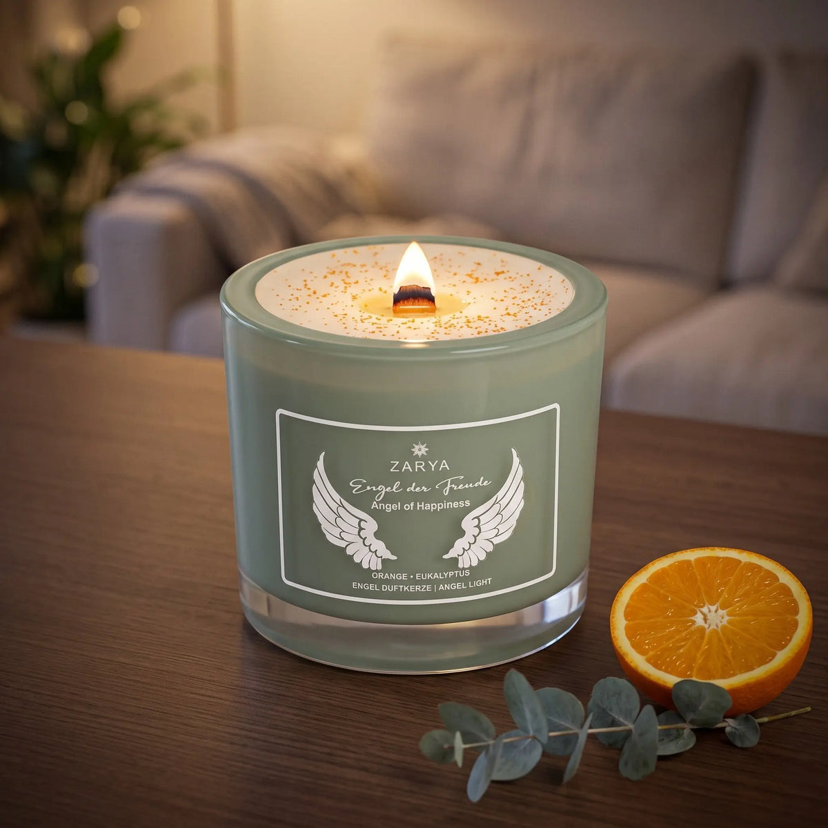 Engelkerze Engel der Freude brennt am Abend auf einem dunklen Holztisch im Wohnzimmer, daneben eine halbe Orange und ein Eukalyptuszweig – Spirit of Gaia