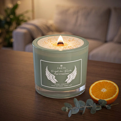 Engelkerze Engel der Freude brennt am Abend auf einem dunklen Holztisch im Wohnzimmer, daneben eine halbe Orange und ein Eukalyptuszweig – Spirit of Gaia