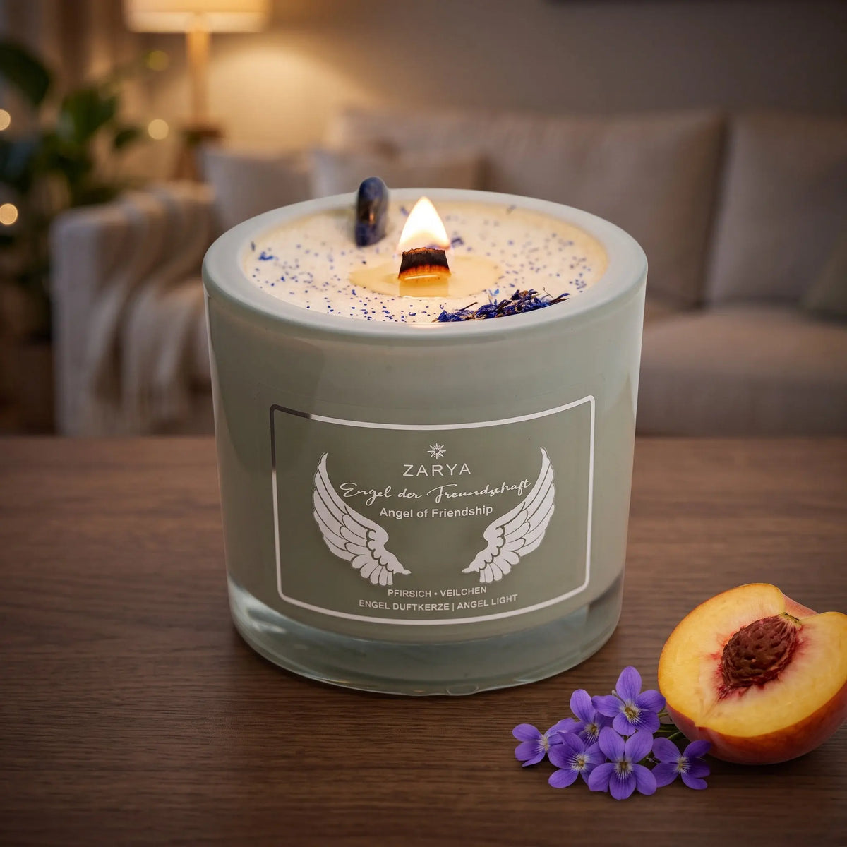 Engelkerze Engel der Freundschaft brennt am Abend auf einem dunklen Holztisch im Wohnzimmer, daneben ein halber Pfirsich und Veilchenblüten – Spirit of Gaia
