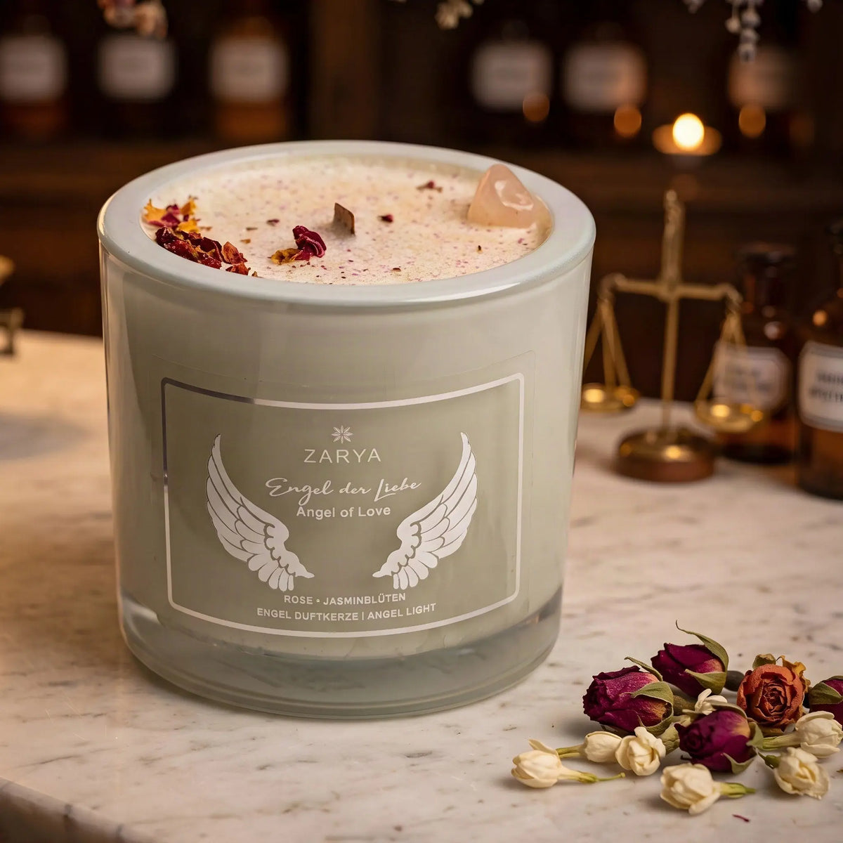 Engelkerze Engel der Liebe von Zarya: salbeigrünes Glas mit weißem Aufdruck auf der Marmortheke einer alten Apotheke am Abend, daneben getrocknete Rosenknospen und Jasminblüten – Spirit of Gaia