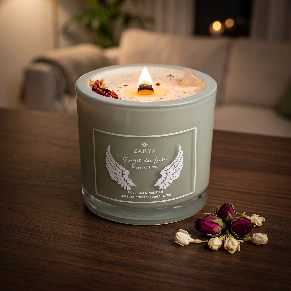 Engelkerze Engel der Liebe brennt am Abend auf einem dunklen Holztisch im Wohnzimmer, daneben getrocknete Rosenknospen und Jasminblüten – Spirit of Gaia