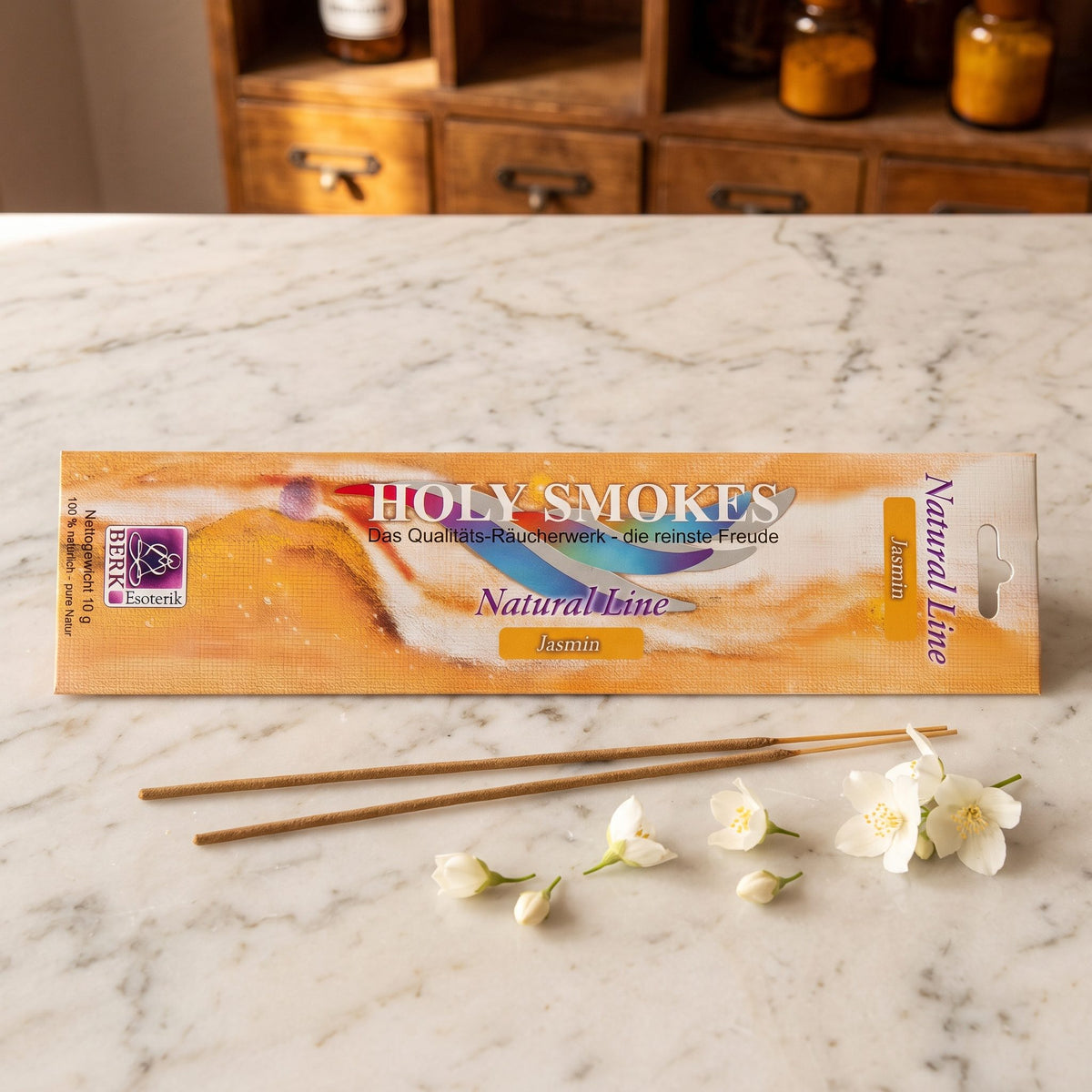 Jasmin Räucherstäbchen der Holy Smokes Natural Line von Berk: flache Packung auf einer Marmortheke vor einem alten Holzschrank mit Schubladen und Bernsteinglasflaschen, davor zwei lose Stäbchen und weiße Jasminblüten