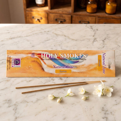 Jasmin Räucherstäbchen der Holy Smokes Natural Line von Berk: flache Packung auf einer Marmortheke vor einem alten Holzschrank mit Schubladen und Bernsteinglasflaschen, davor zwei lose Stäbchen und weiße Jasminblüten