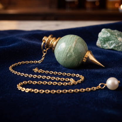Aventurin-Kugelpendel mit vergoldeter Messingspitze, ca. 3 cm: liegt mit locker gelegter Kette auf dunkelblauem Samt, daneben ein Stück roher Aventurin, abends auf der Theke einer alten Apotheke – Spirit of Gaia