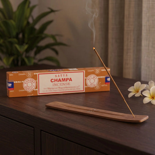 Satya Champa Räucherstäbchen: glimmendes Räucherstäbchen mit feinem Rauch im Holzhalter, dahinter die Packung, auf einem Sideboard am Abend