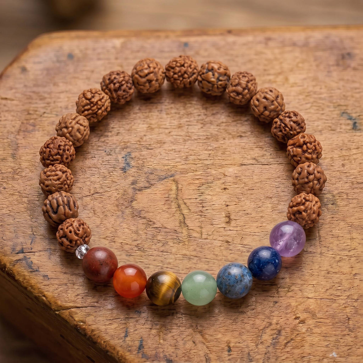 Draufsicht auf das Chakra Rudraksha Armband auf altem Holz: sieben Steine von Rotbraun bis Violett zwischen zerfurchten Rudraksha-Samen