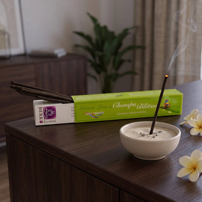 Champa-Blüten Räucherstäbchen naturrein, Holy Smokes Green Line: glimmendes Räucherstäbchen in einer Keramikschale mit Sand, dahinter die geöffnete Packung, auf einem Sideboard am Abend
