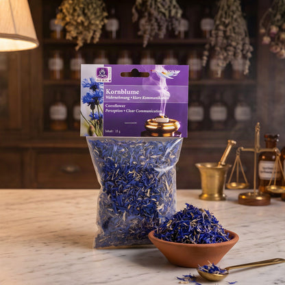 Kornblume von Berk: Tüte mit violettem Kartonkopf und blauen Blütenblättern auf der Marmortheke einer Apotheke am Abend, daneben eine Terracottaschale mit Kornblumenblüten und ein Messinglöffel