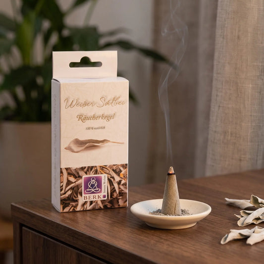 Räucherpyramiden Weißer Salbei von Berk: glimmender Räucherkegel mit feinem Rauch auf einem Keramiktellerchen, dahinter die Schachtel, auf einem Sideboard am Abend