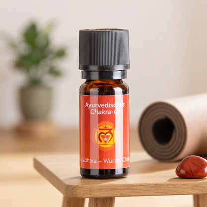 Ayurvedisches Chakra-Öl Muladhara (Wurzel-Chakra): 10-ml-Fläschchen auf einem Holzhocker neben einer gerollten Yogamatte, daneben roter Jaspis