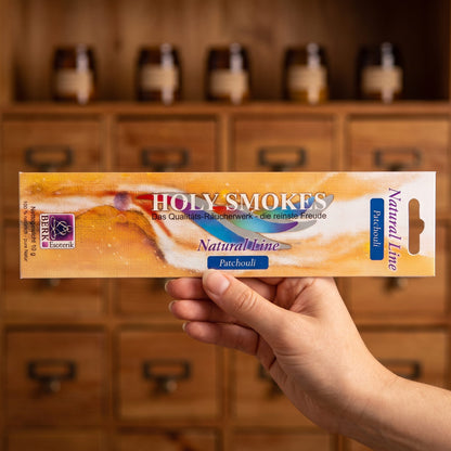 Eine Hand hält die flache Packung Patchouli Natural Line von Holy Smokes, etwa 24 cm lang, vor einem Holzschrank mit Schubladen