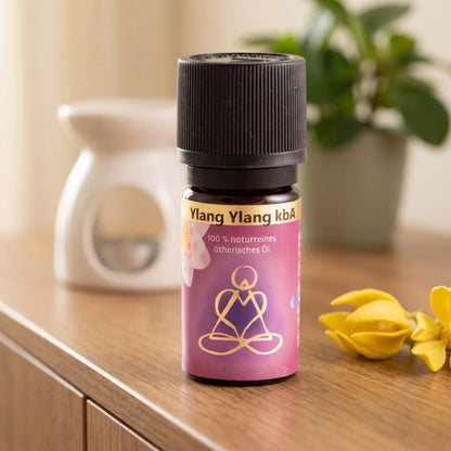 Ätherisches Öl Ylang Ylang: Fläschchen auf einem Holz-Sideboard im Wohnzimmer, im Hintergrund eine weiße Duftlampe, daneben Ylang-Ylang-Blüten