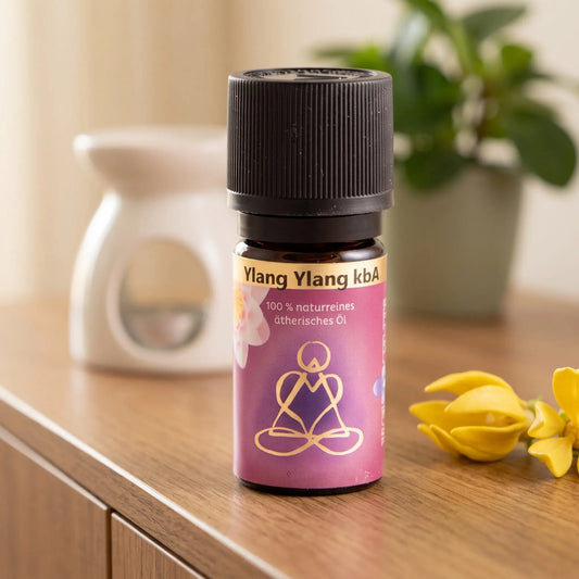 Ätherisches Öl Ylang Ylang: Fläschchen auf einem Holz-Sideboard im Wohnzimmer, im Hintergrund eine weiße Duftlampe, daneben Ylang-Ylang-Blüten