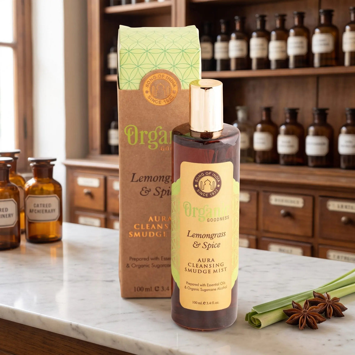 Aura Reinigungsspray Zitronengras und Gewürze von Organic Goodness: braune Sprühflasche mit goldener Kappe neben der Kraftpapier-Schachtel auf einer Marmortheke vor einem Apothekerschrank