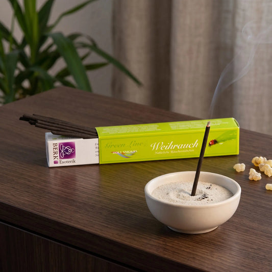 Weihrauch Räucherstäbchen naturrein, Holy Smokes Green Line: glimmendes Räucherstäbchen in einer Keramikschale mit Sand, dahinter die geöffnete Packung, auf einem Sideboard am Abend
