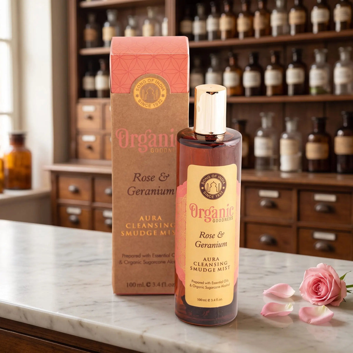 Aura Reinigungsspray Rose und Geranie von Organic Goodness: braune Sprühflasche mit goldener Kappe neben der Kraftpapier-Schachtel auf einer Marmortheke vor einem Apothekerschrank