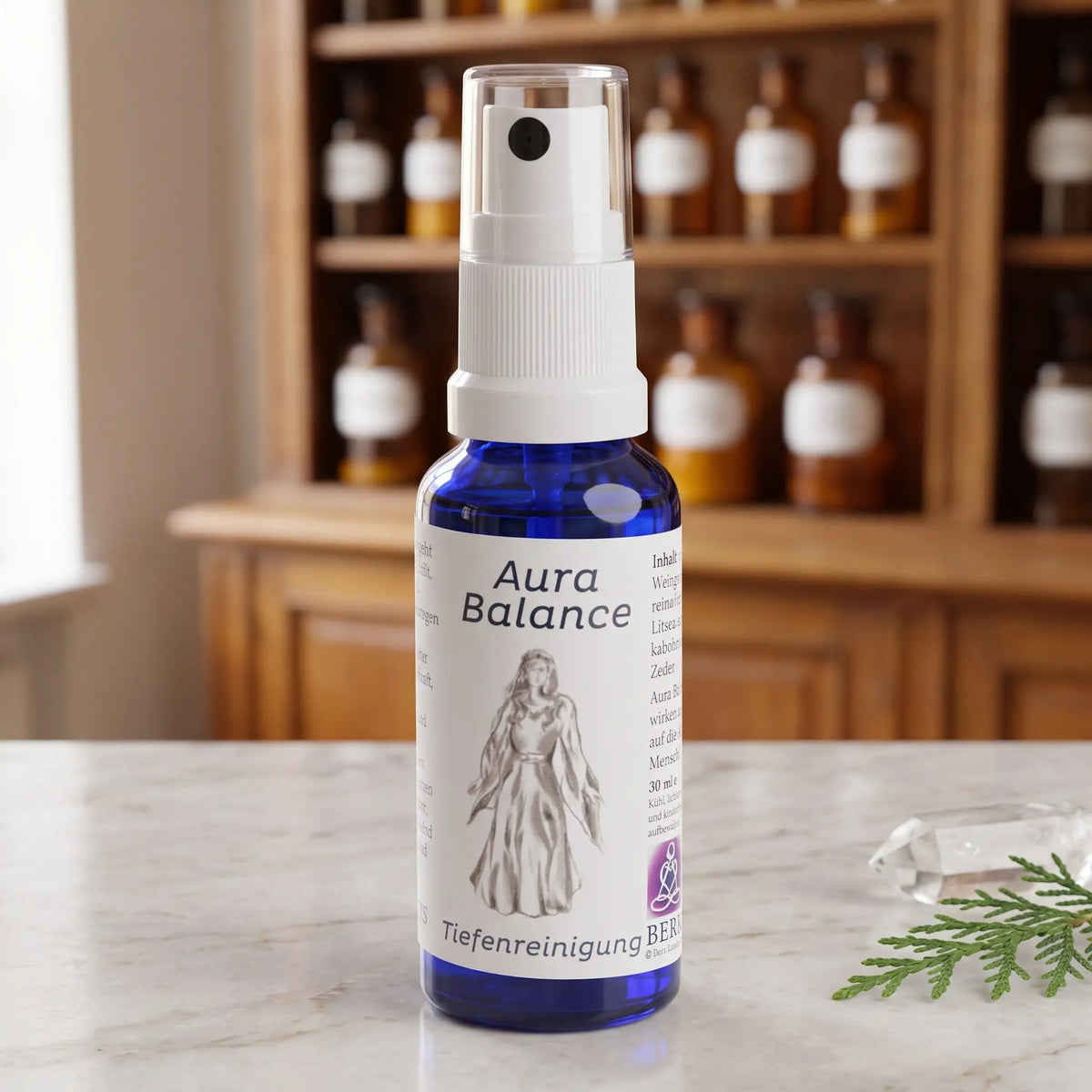 Energiespray Tiefenreinigung (Aura Balance): blaue Sprühflasche mit weißem Etikett auf Marmortheke vor Apothekerschrank, daneben ein Bergkristall und ein Zedernzweig – Spirit of Gaia