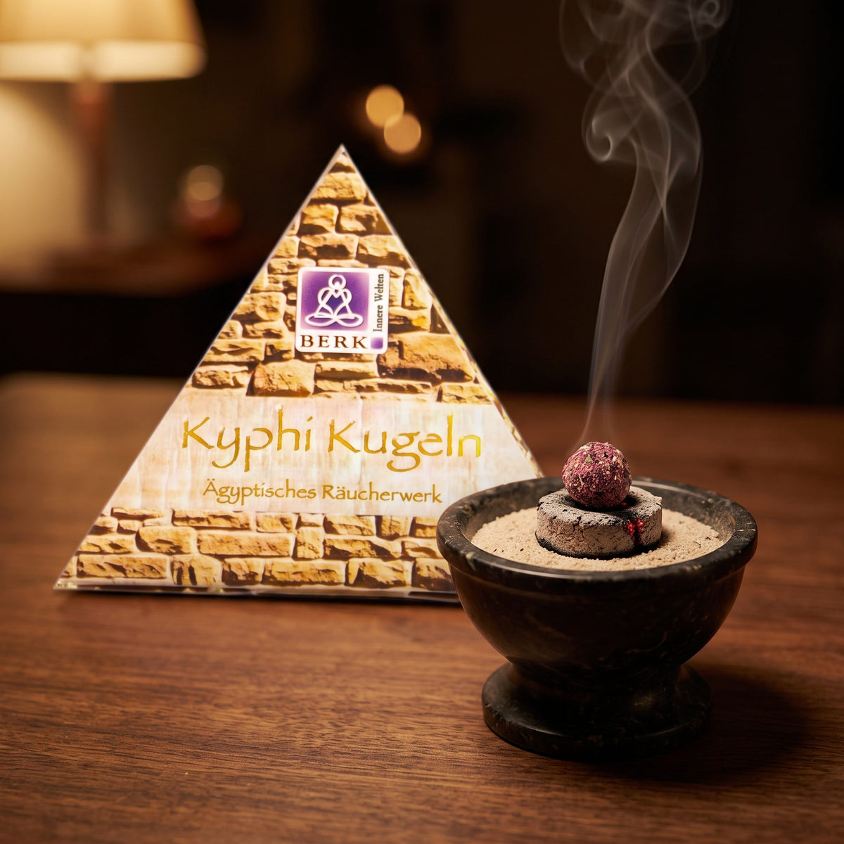 Kyphi Kugeln von Berk: eine Kyphi-Kugel glimmt auf der Räucherkohle in einer Specksteinschale, dahinter die Pyramidenverpackung, feiner Rauch steigt am Abend auf – Spirit of Gaia