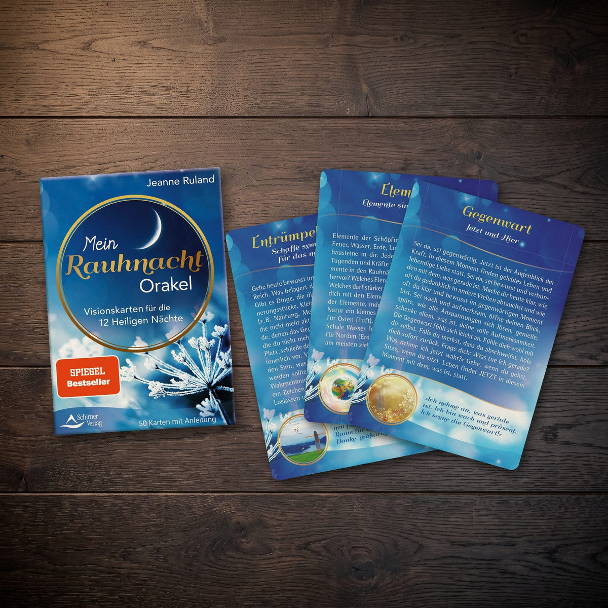 Drei Karten des Rauhnacht-Orakels mit den Titeln Gegenwart, Elemente und Entrümpeln und Lösen liegen neben der blauen Schachtel auf einem Holztisch