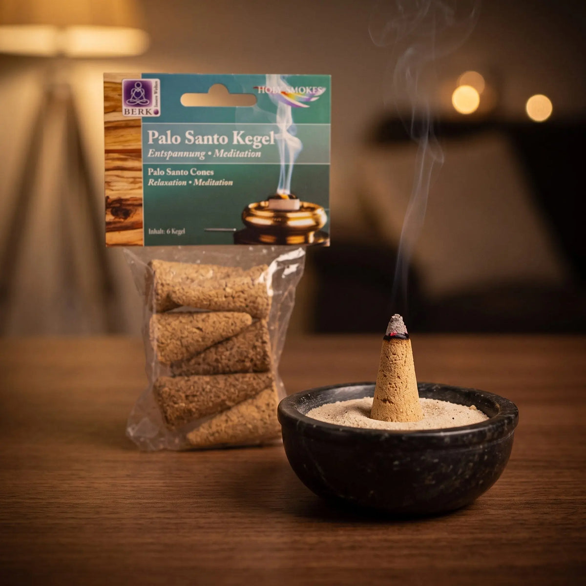 Ein Palo-Santo-Kegel glimmt auf Sand in einer Specksteinschale, dahinter die Tüte, feiner Rauch steigt auf