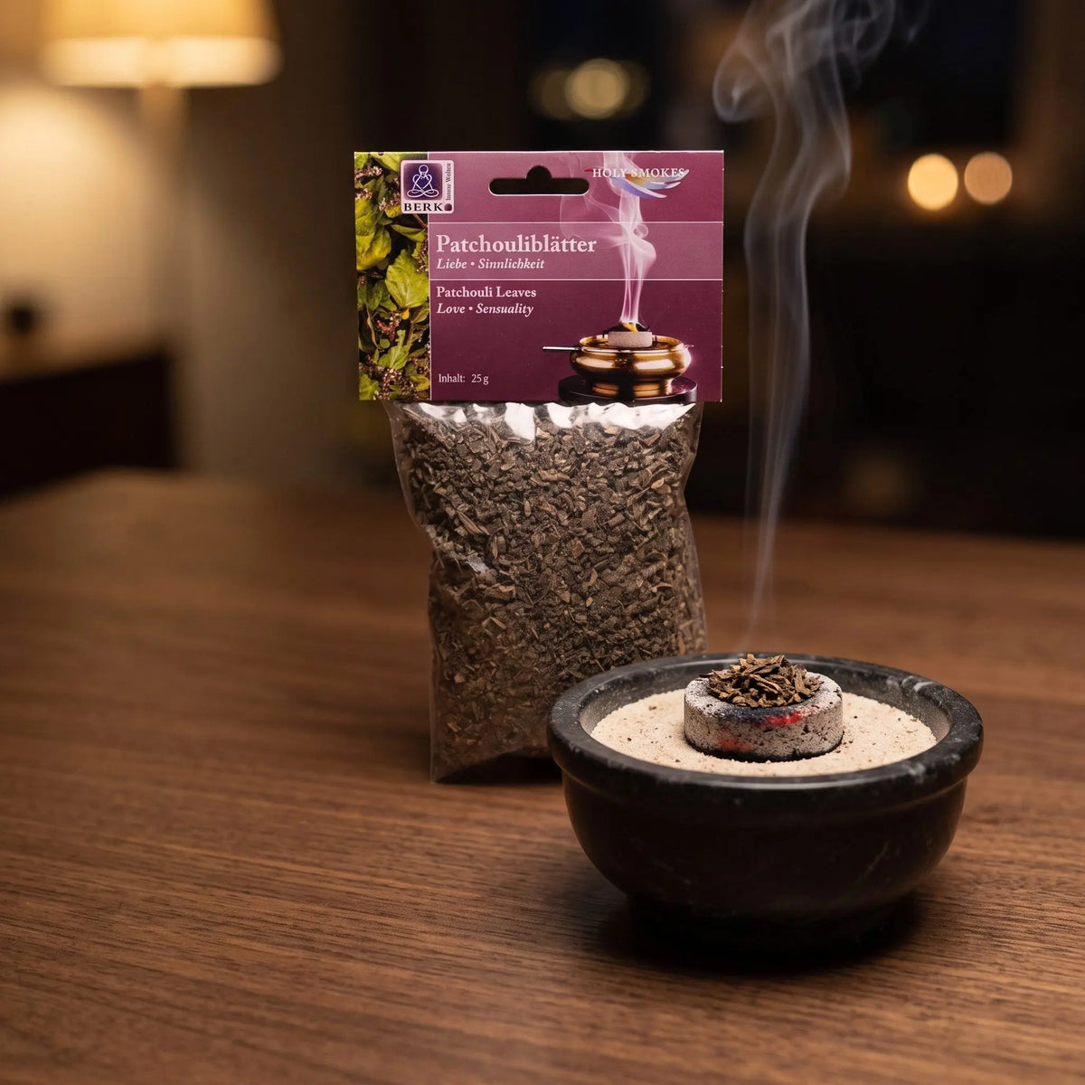 Patchouliblätter, sortenreines Räucherwerk von Berk, 25 g: eine Prise Patchouliblätter glimmt auf der Räucherkohle, dahinter die Tüte, feiner Rauch steigt auf einer dunklen Holzplatte am Abend auf – Spirit of Gaia
