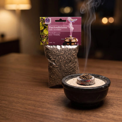 Patchouliblätter, sortenreines Räucherwerk von Berk, 25 g: eine Prise Patchouliblätter glimmt auf der Räucherkohle, dahinter die Tüte, feiner Rauch steigt auf einer dunklen Holzplatte am Abend auf – Spirit of Gaia