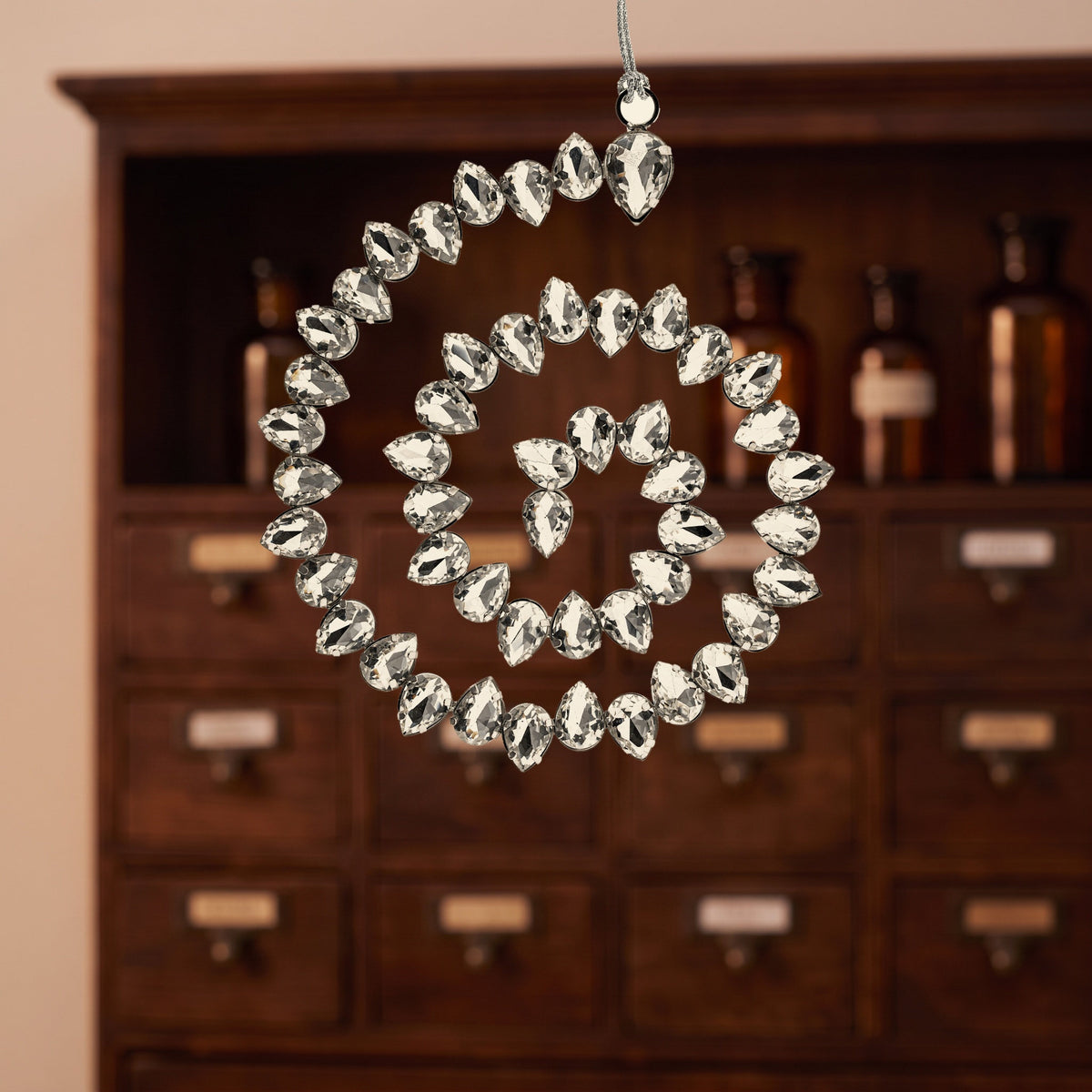 Kristallspirale aus facettierten Glassteinen in silbernem Stahl hängt an der Schlaufe vor einem dunklen Holzschrank mit kleinen Schubladen und braunen Glasflaschen